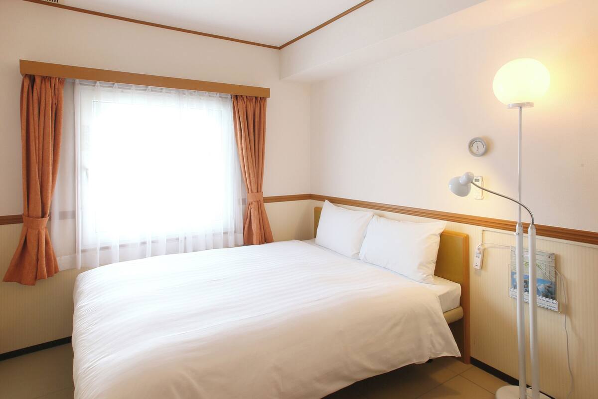 A hotel room with a bed, curtains, a bedside lamp, a clock, and a thermostat on the wall.