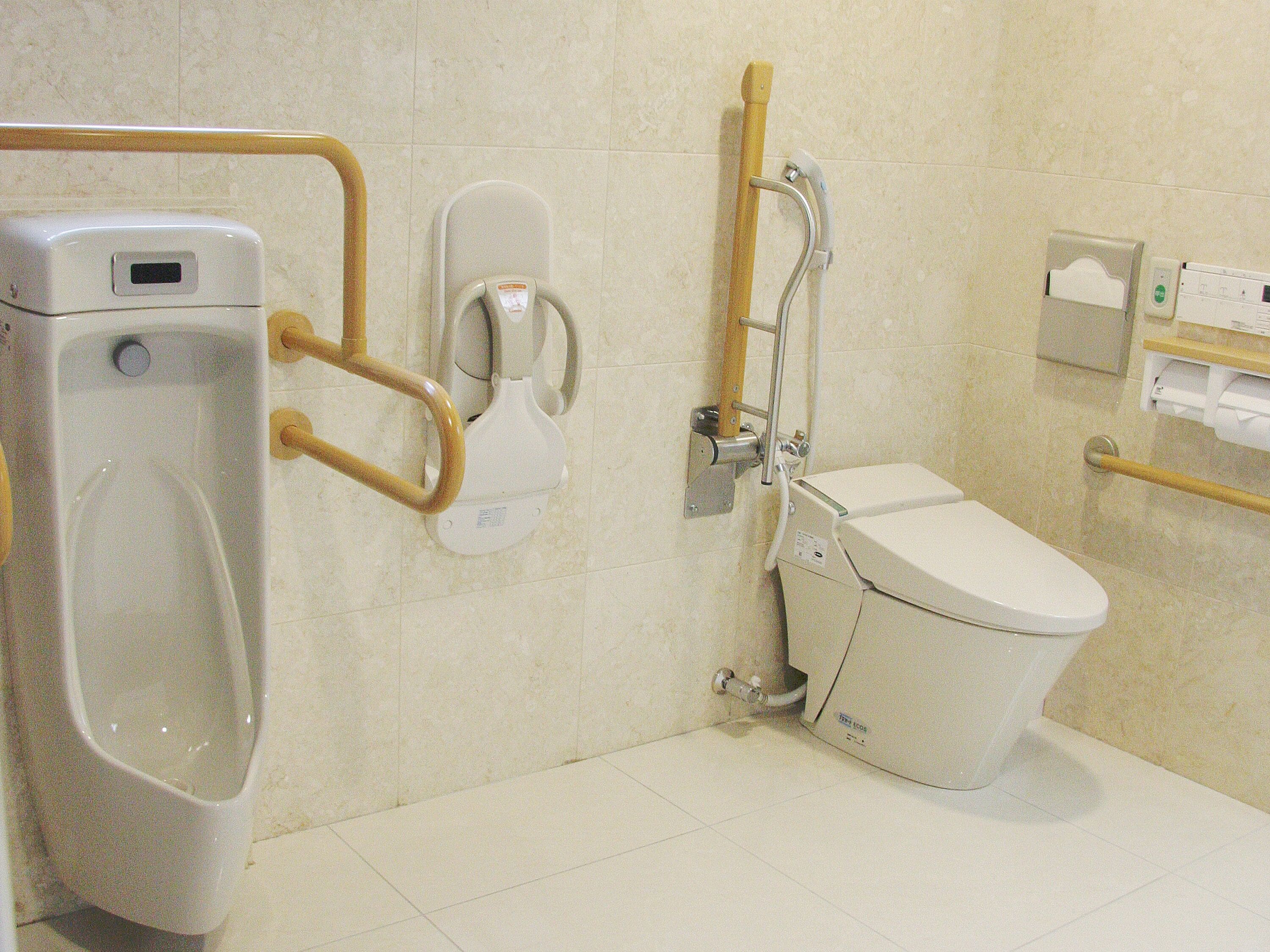 Une salle de bain adaptée aux personnes handicapées, équipée de toilettes, d’un bidet et d’un urinoir.