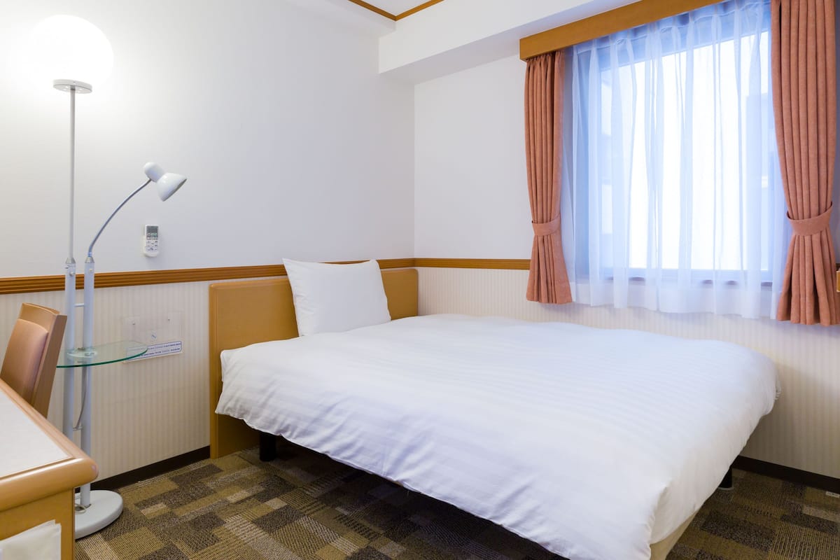A hotel room with a bed, a desk, a lamp, and a window with curtains.