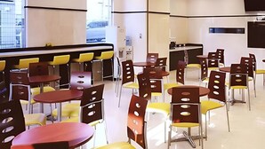 Un área de comedor moderna con varias mesas y sillas, un reloj a la vista y una cocina al fondo.