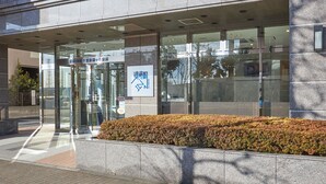 A modern building entrance with glass doors, a sign in a foreign language, and a landscaped area with bushes.