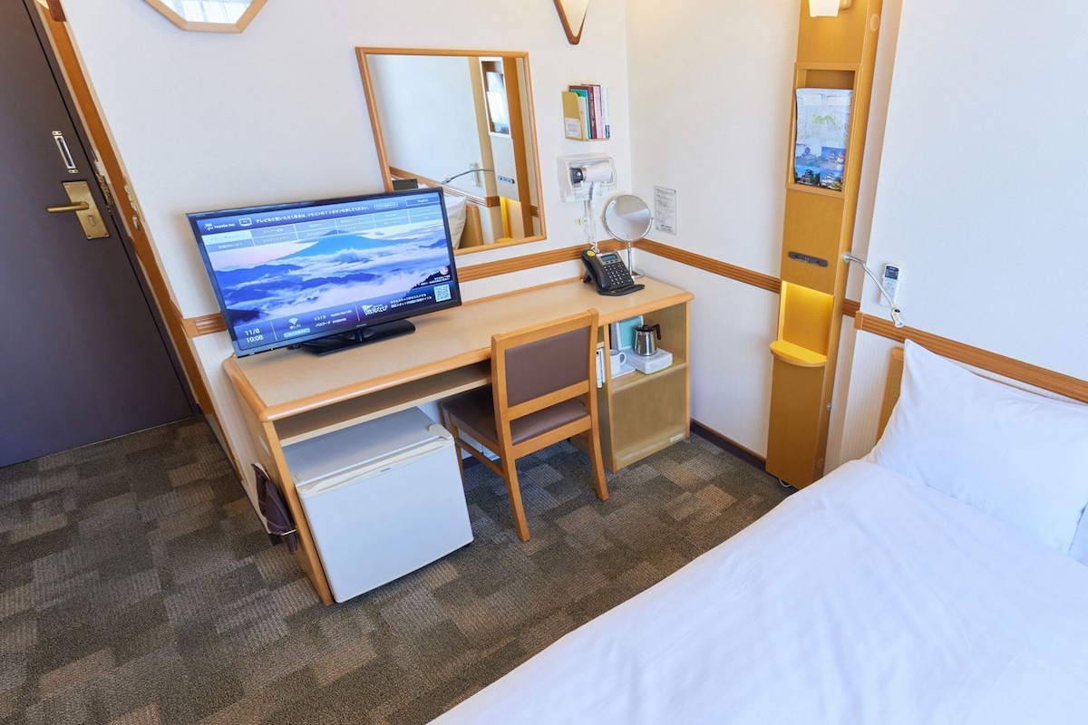 A hotel room with a bed, a desk with a computer, a small fridge, a mirror, and a bedside table.