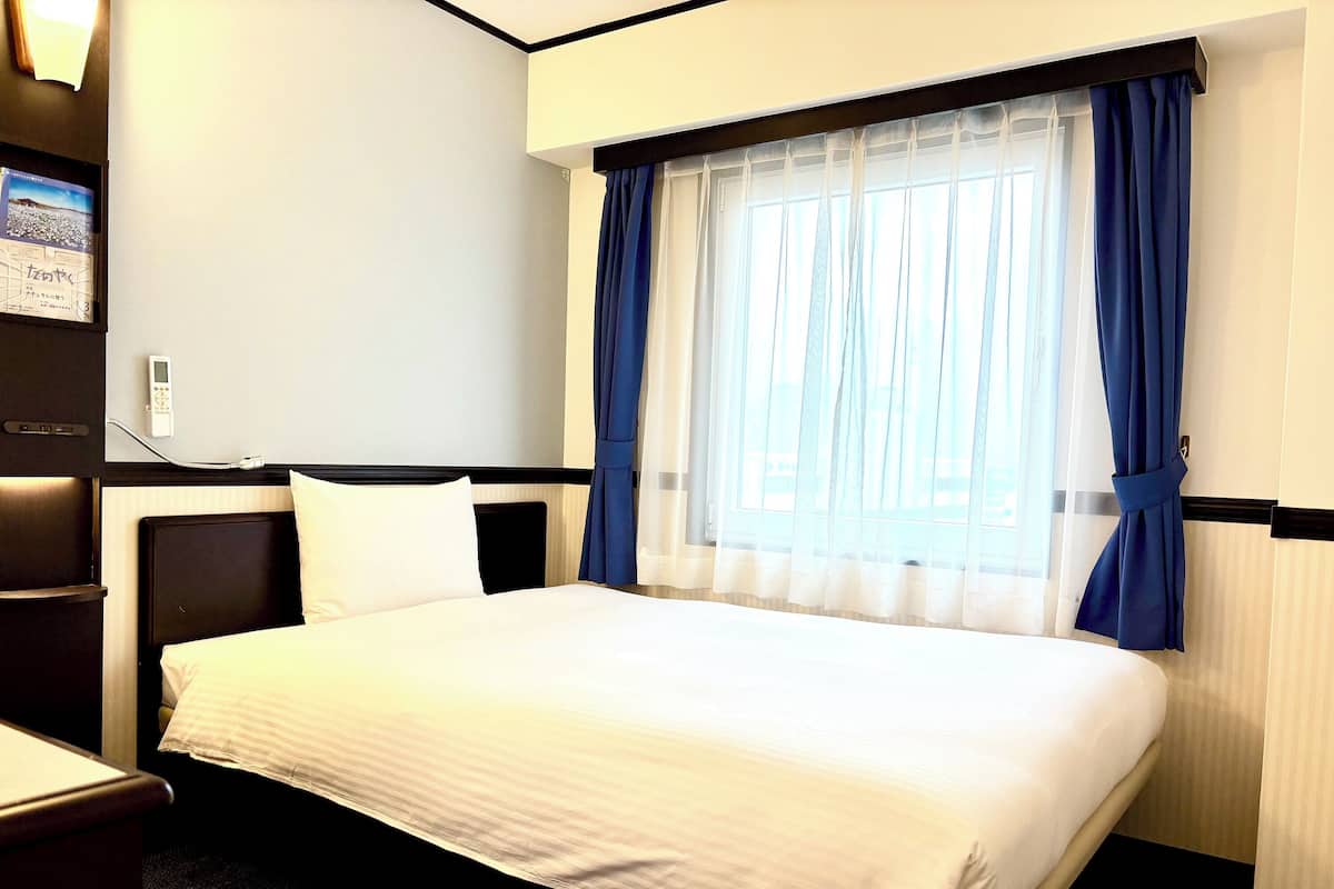A hotel room with a neatly made bed, a bedside table, a lamp, and a window with blue curtains.