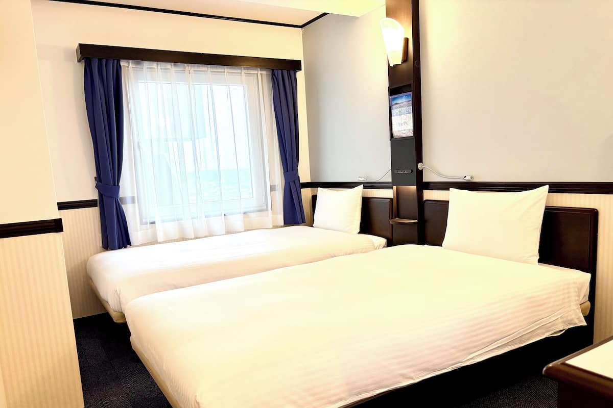A hotel room with a large bed, a bedside table, a nightstand, a window with curtains, and a wall-mounted light.
