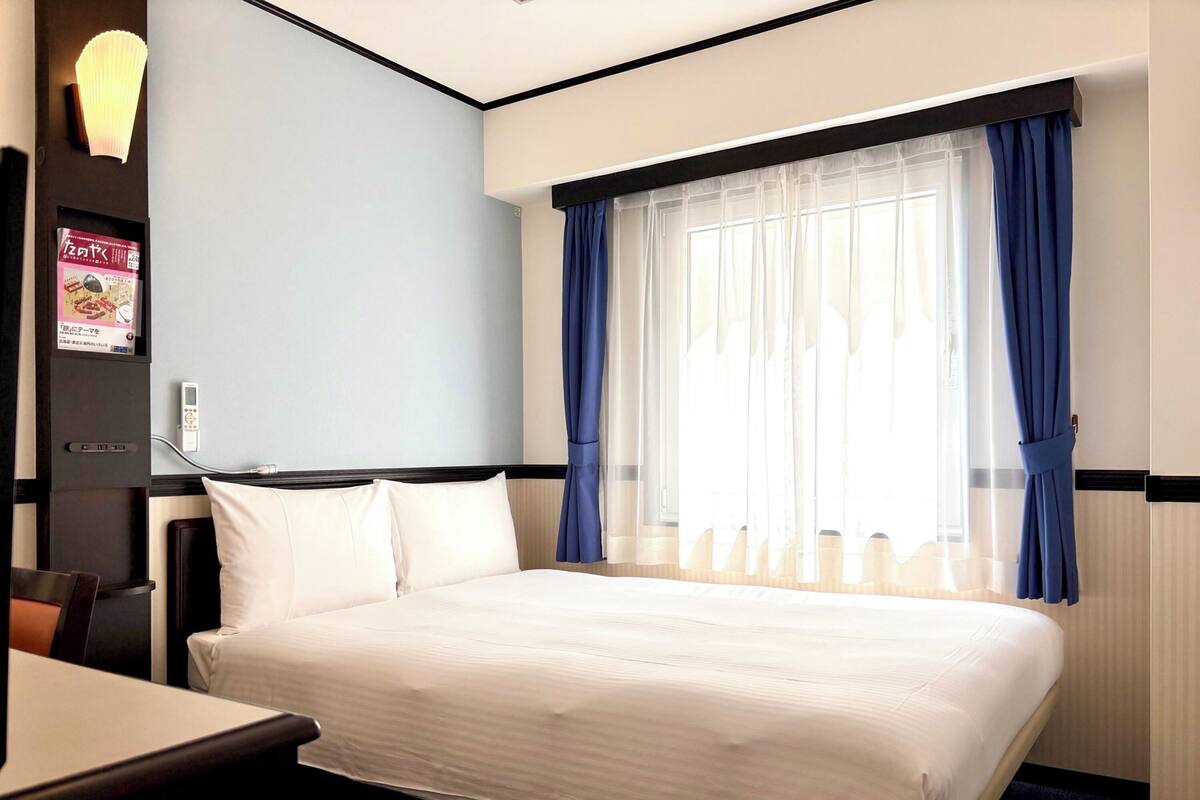 A hotel room with a bed, a desk, a chair, a lamp, and a window with curtains.
