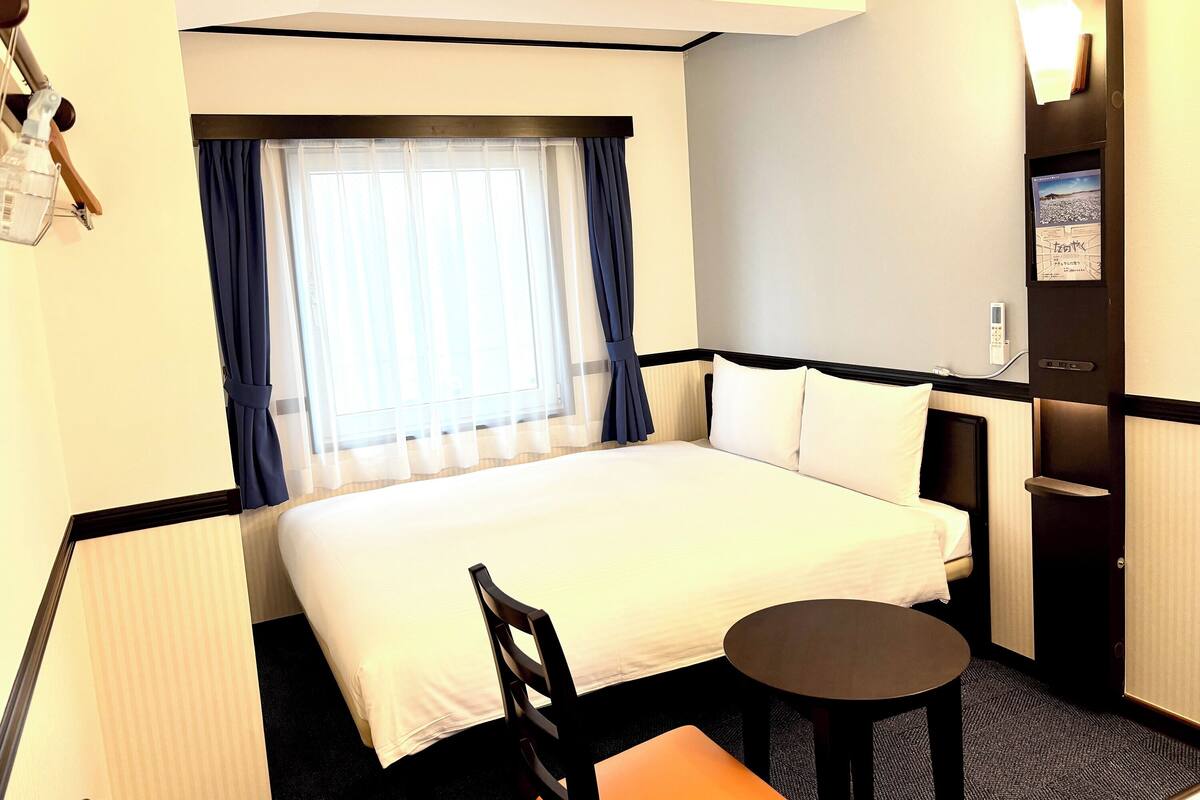 A hotel room with a bed, a small table, a chair, a window with curtains, and a wall-mounted TV.