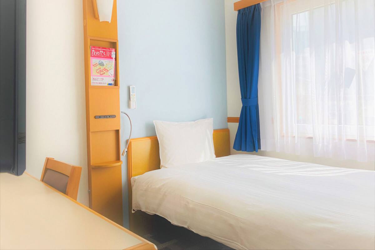 Ein Hotelzimmer mit einem Bett, einem Schreibtisch, einem Stuhl, einem Regal mit einem Buch und einem Fenster mit Vorhängen.