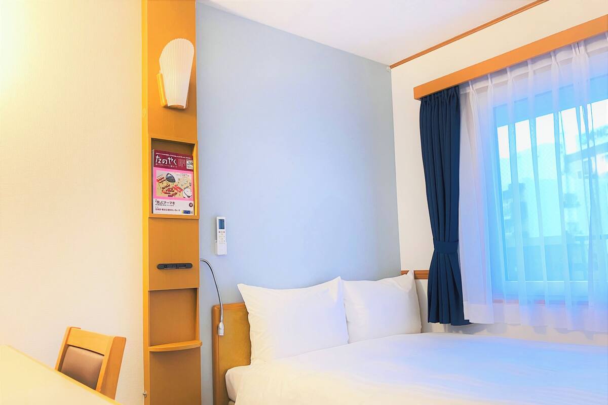 Ein Hotelzimmer mit Bett, Schreibtisch, Stuhl, Nachttisch, Wandlampe, einem Fenster mit Vorhängen und einem Poster an der Wand.