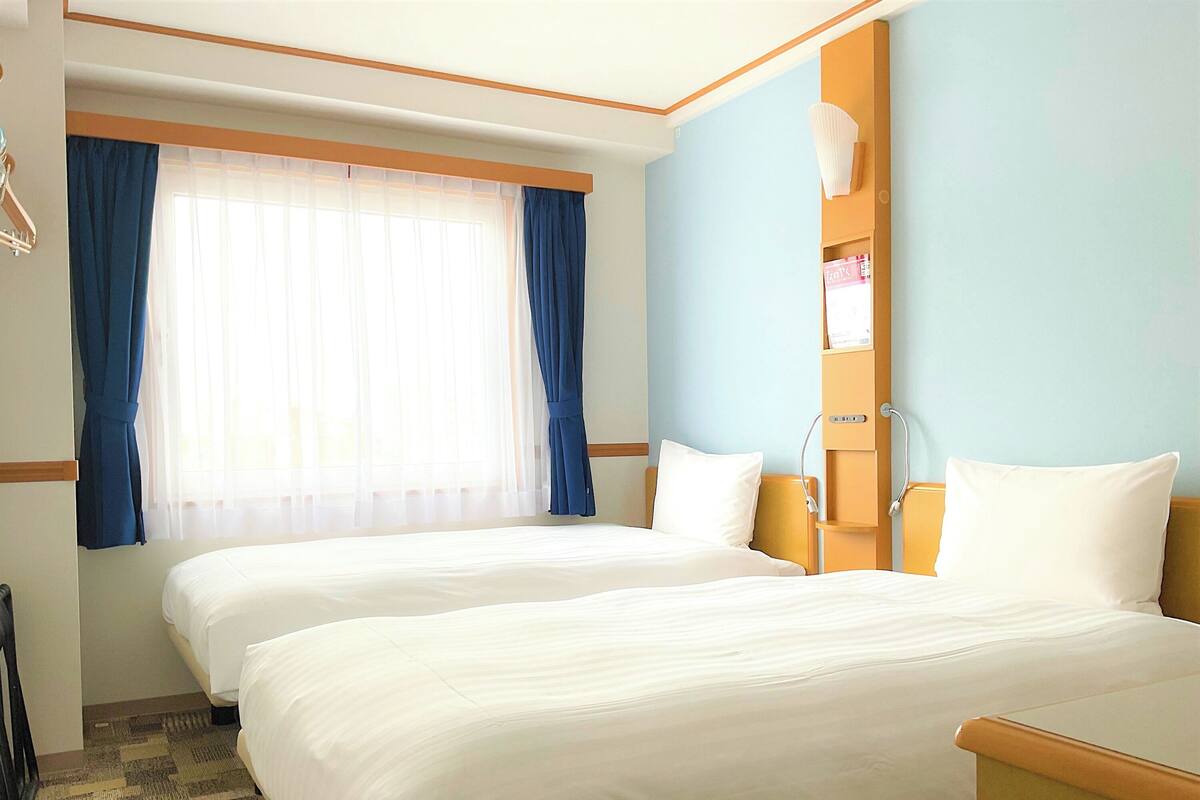Ein Hotelzimmer mit zwei Betten, einem Fenster mit blauen Vorhängen, einem Nachttisch mit Lampe und einem hölzernen Kopfteil.