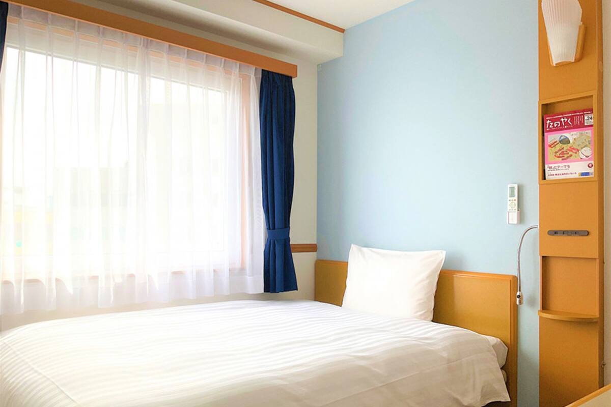 Ein Hotelzimmer mit einem Bett, einem Fenster mit durchsichtigen Vorhängen, einem blauen Vorhang, einem weißen Kopfkissen, einem hölzernen Kopfteil, einem Nachttisch und einem an der Wand befestigten Regal mit einer Zeitschrift.