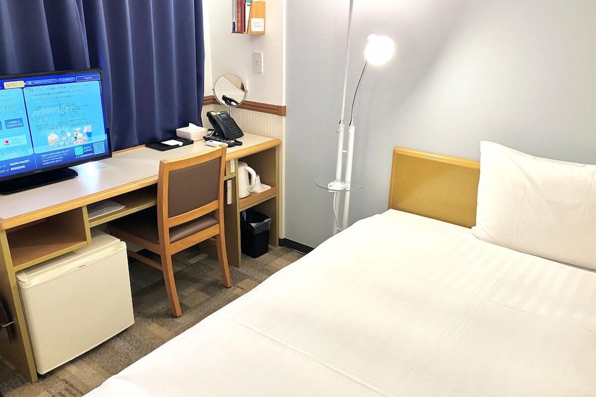 A hotel room with a bed, desk, chair, computer, and a bedside lamp.