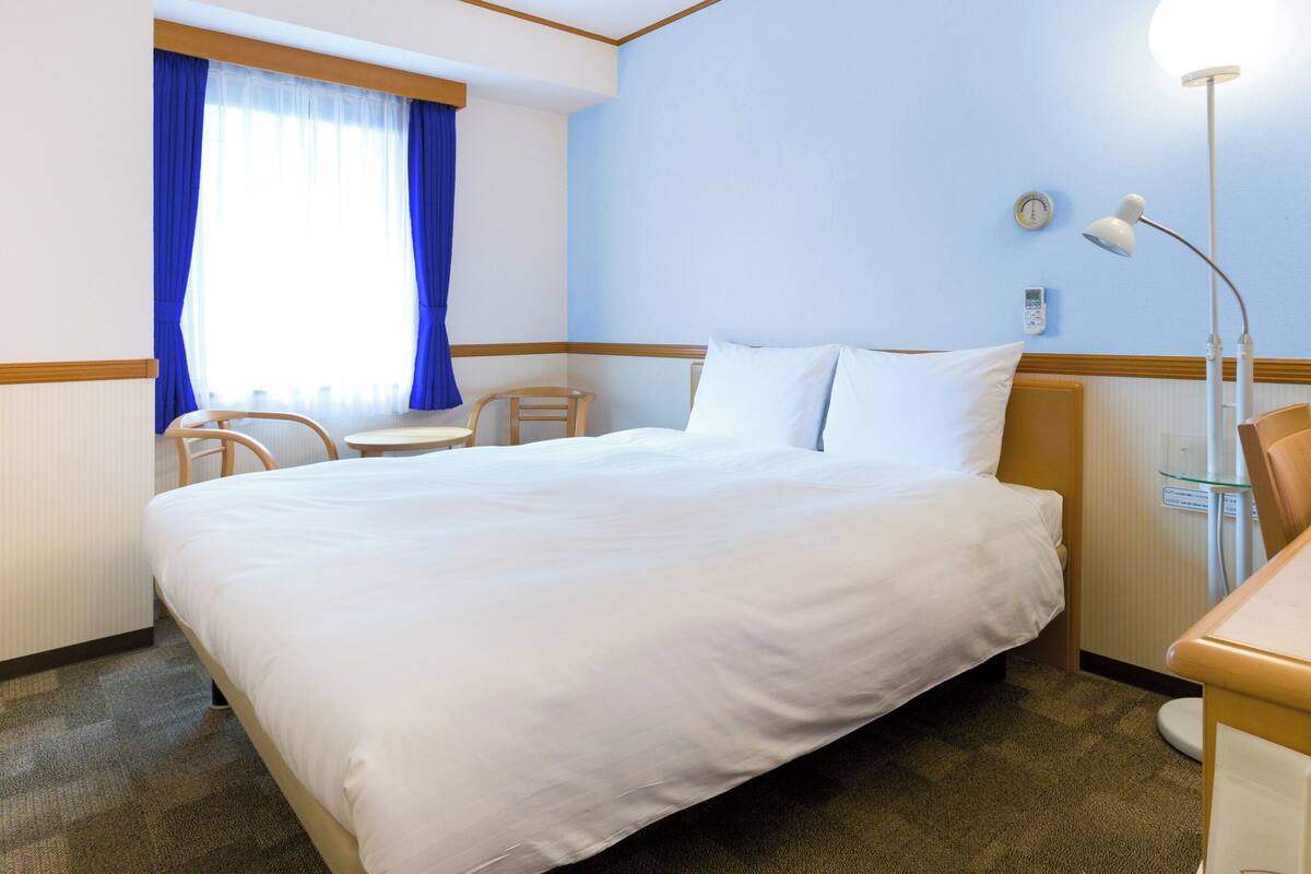A hotel room with a large bed, a desk, a chair, a lamp, and a window with blue curtains.
