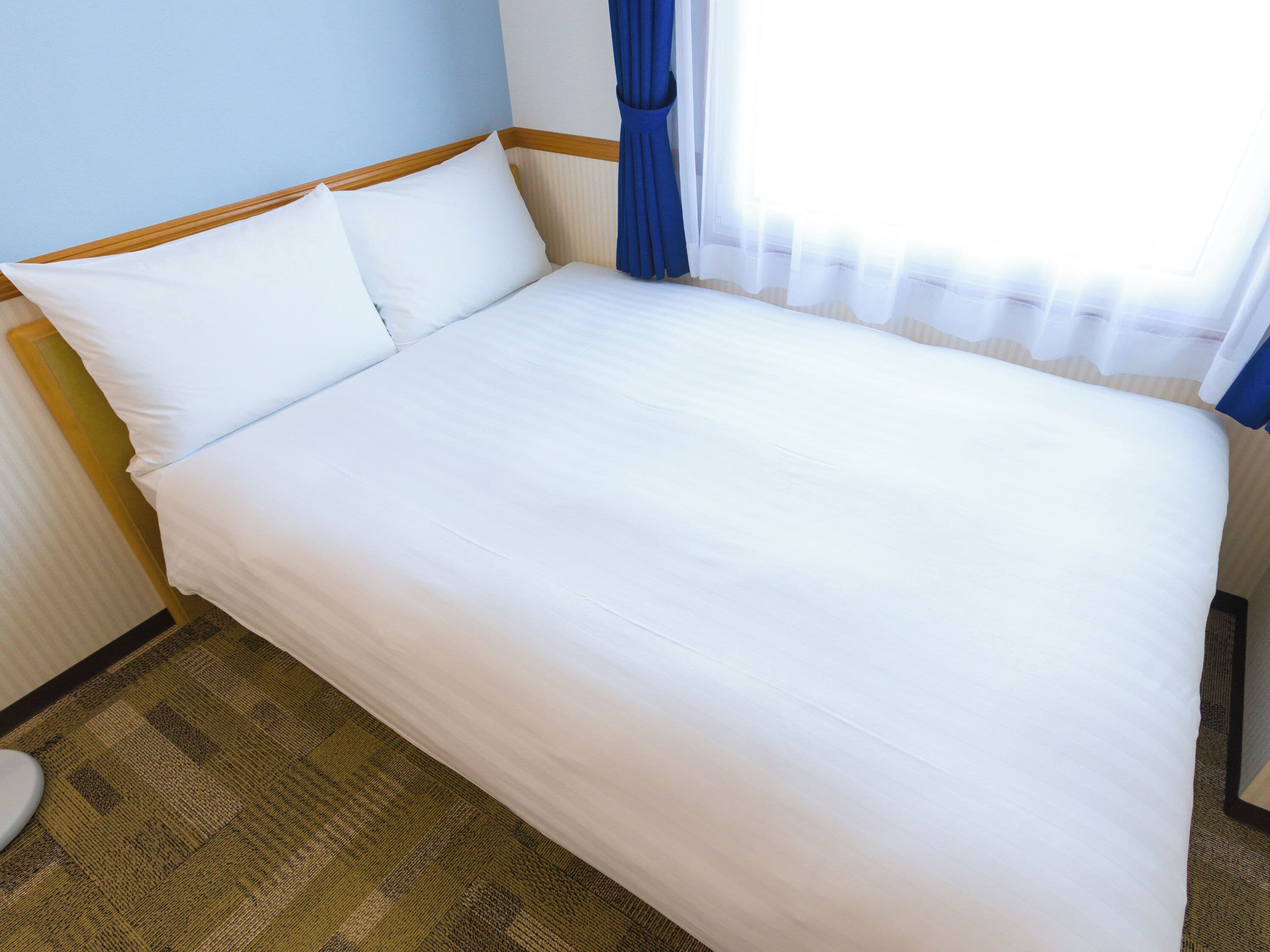 A neatly made bed with white linens in a hotel room with a wooden headboard, a patterned carpet, and a window with blue curtains.
