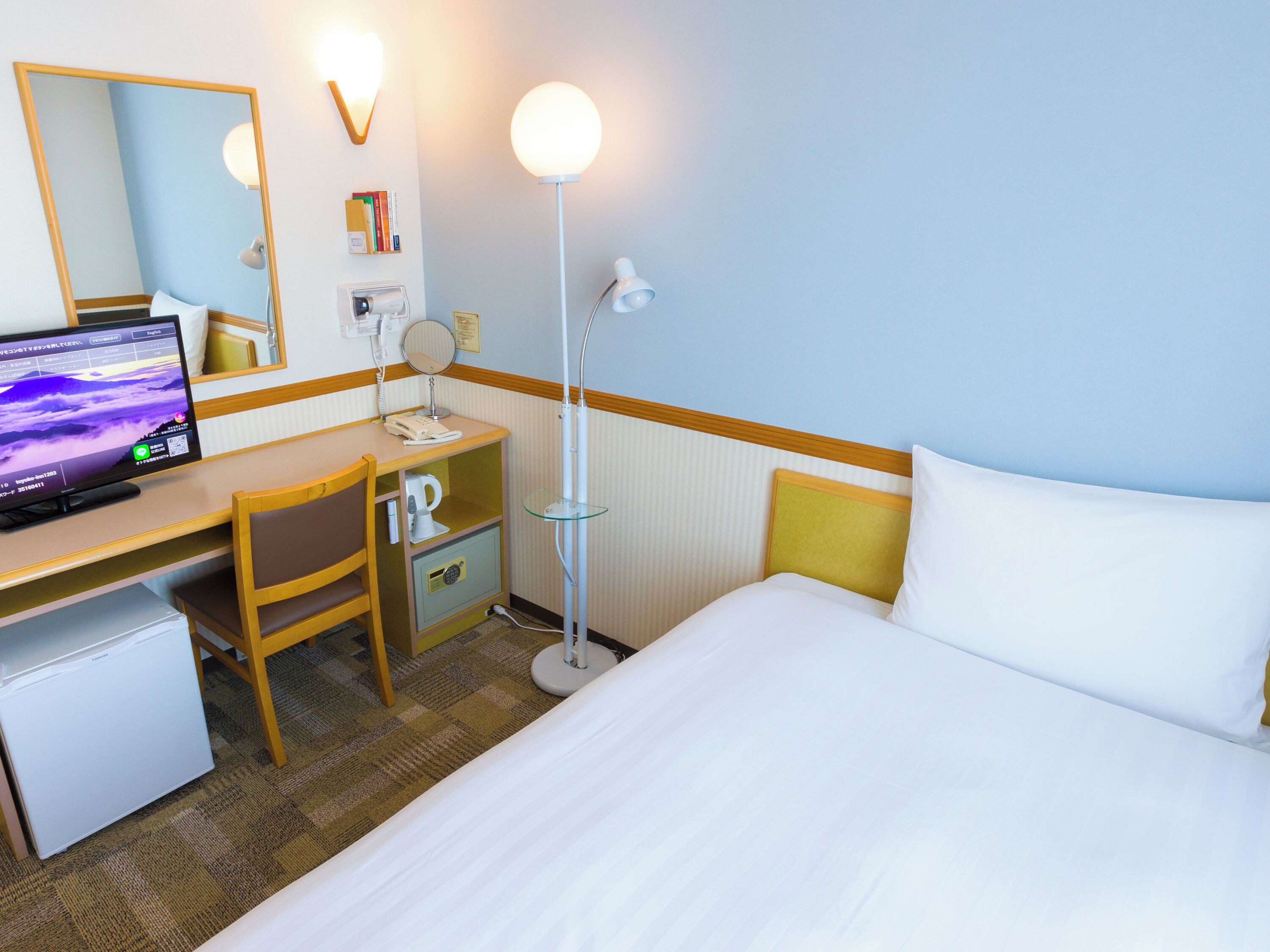 A hotel room with a bed, a desk with a computer, a chair, a desk lamp, a telephone, and a mirror.