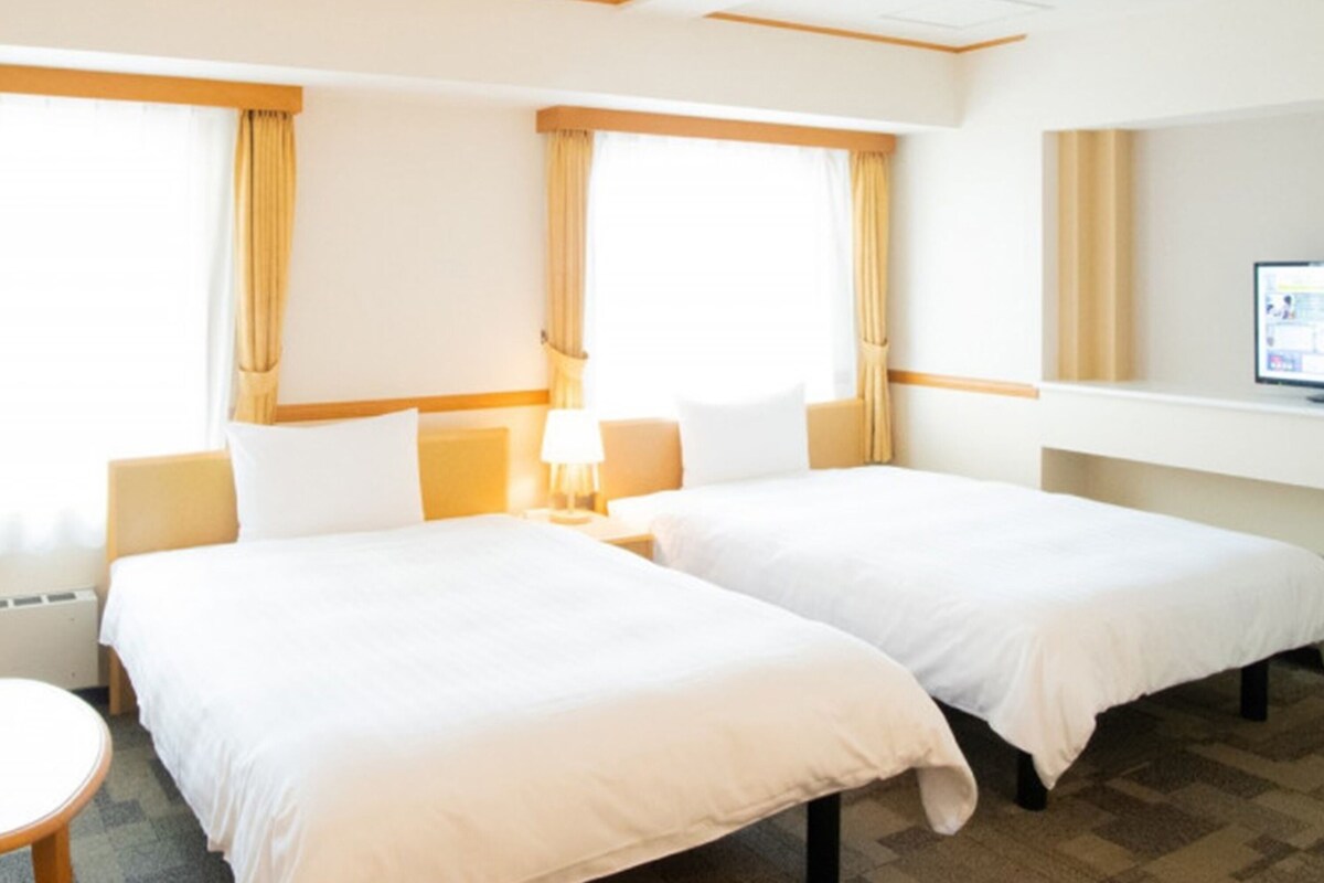 A hotel room with two beds, a small table, a TV, and a window with curtains.