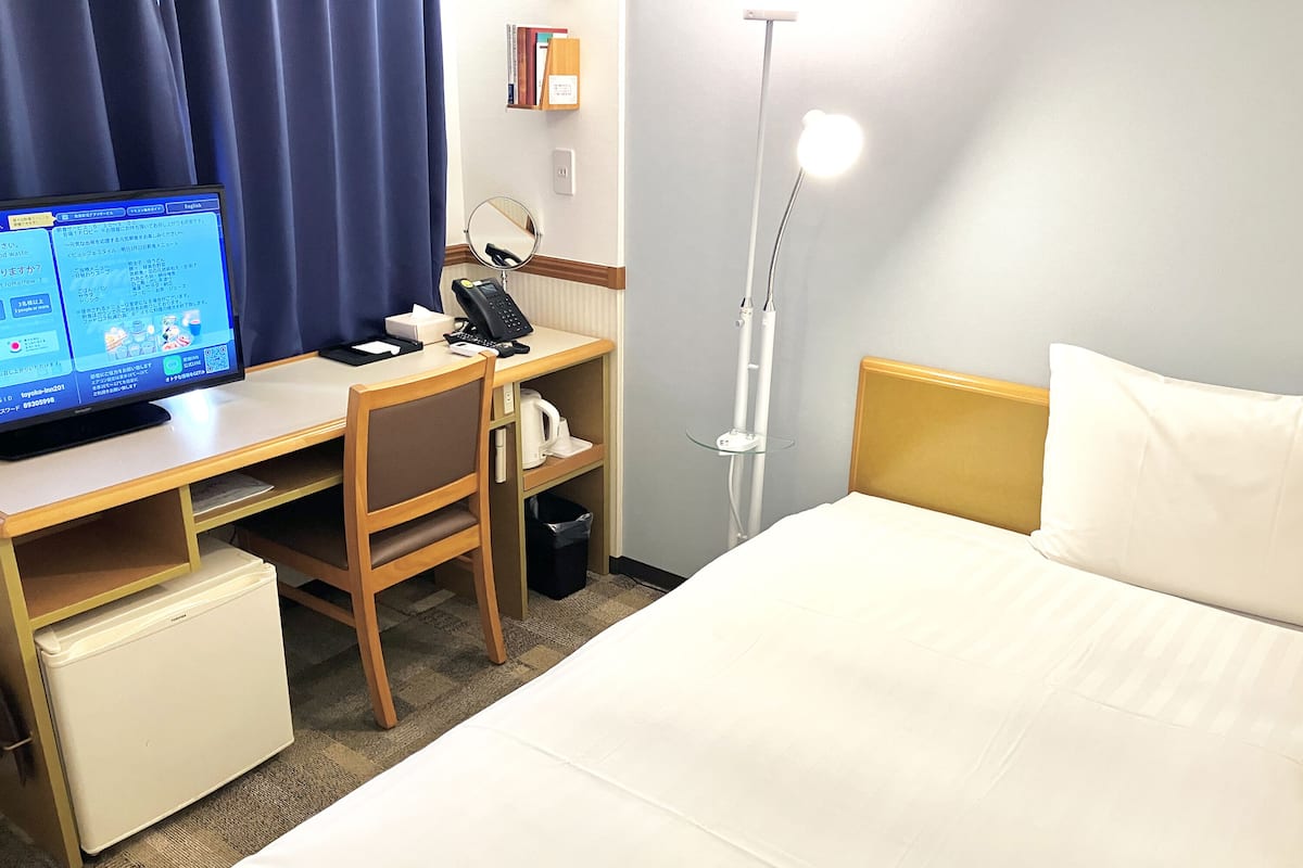 A hotel room with a bed, desk, chair, and a small refrigerator.