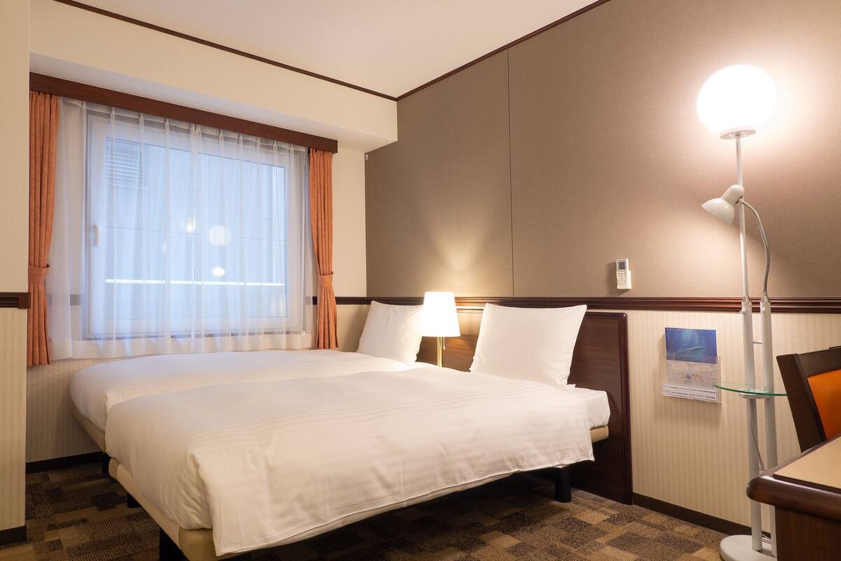 Ein Hotelzimmer mit einem großen Bett, einer Nachttischlampe, einem Fenster mit Vorhängen und einem an der Wand befestigten Telefon.