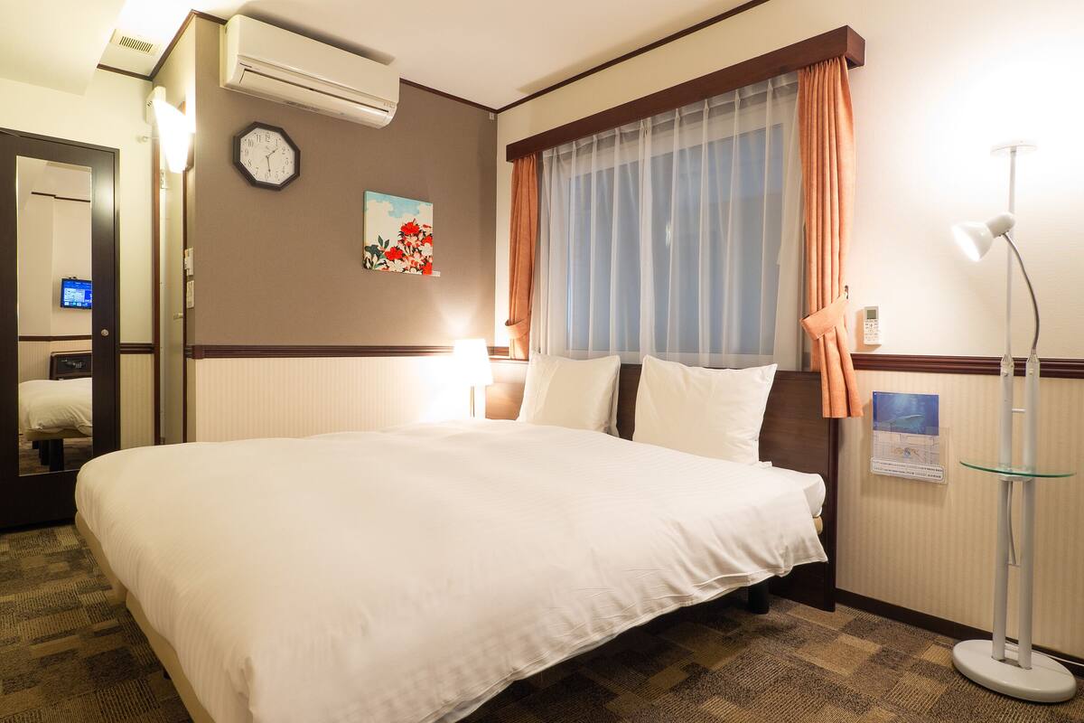 Ein Hotelzimmer mit einem großen Bett, einer Wanduhr, einem Gemälde, einem Fenster mit Vorhängen und einer Stehlampe.