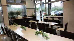 Un restaurant doté de nombreuses tables et chaises, de grandes fenêtres et de plantes d’intérieur.