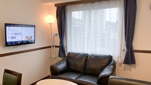 Une chambre d’hôtel avec un canapé en cuir, une petite table ronde, un téléviseur fixé au mur et une fenêtre avec des rideaux.