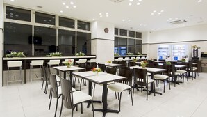 Eine großzügige Cafeteria mit mehreren Tischen, Stühlen, großen Fenstern und Verkaufsautomaten.