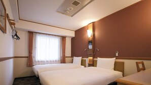 Quarto de hotel com duas camas, uma mesa, uma cadeira, mesa de cabeceira, abajur, telefone e lixeira.