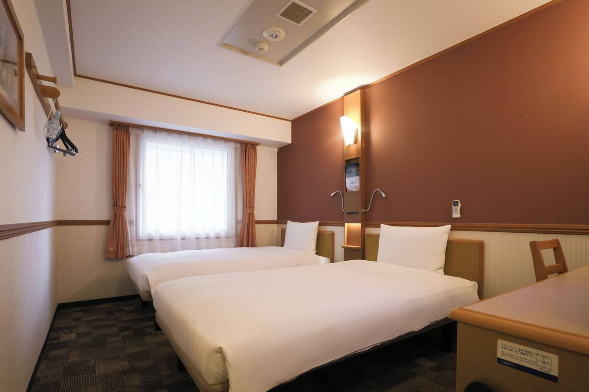 A hotel room with two beds, a desk, a chair, a bedside table, a lamp, a telephone, and a trash bin.