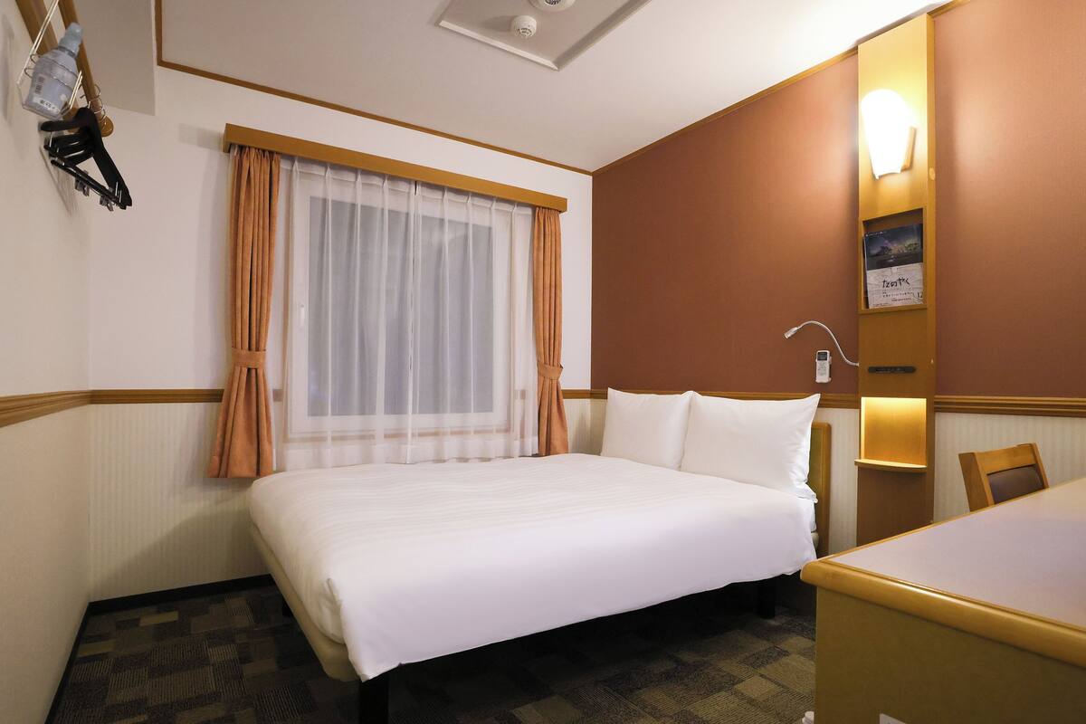 A hotel room with a bed, a desk, a chair, a window with curtains, a wall-mounted lamp, and a small shelf with a picture.