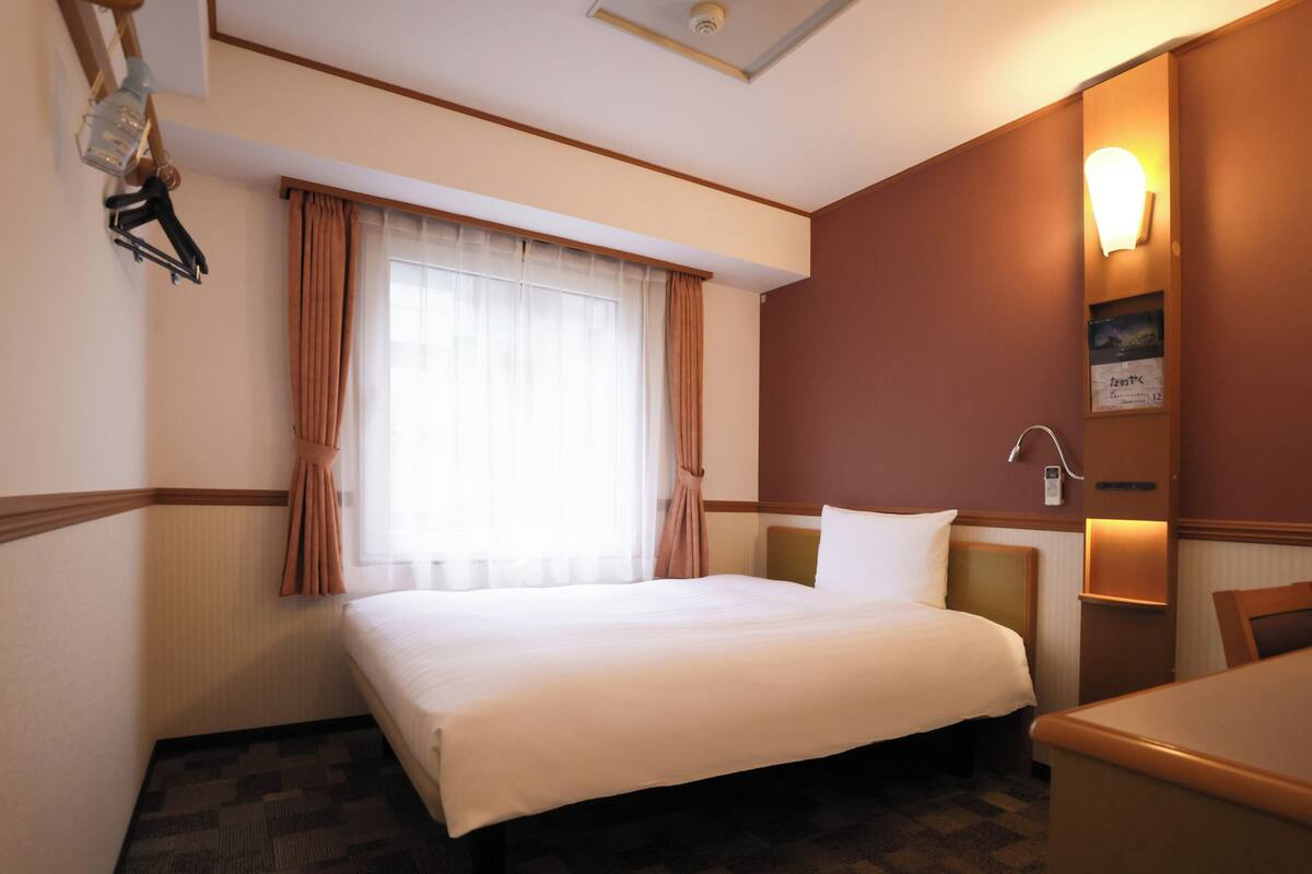 A hotel room with a bed, a desk, a chair, a window with curtains, a wall-mounted lamp, and a clock.