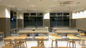Uma sala de aula com mesas e cadeiras dispostas em fileiras, janelas amplas e luminárias no teto.