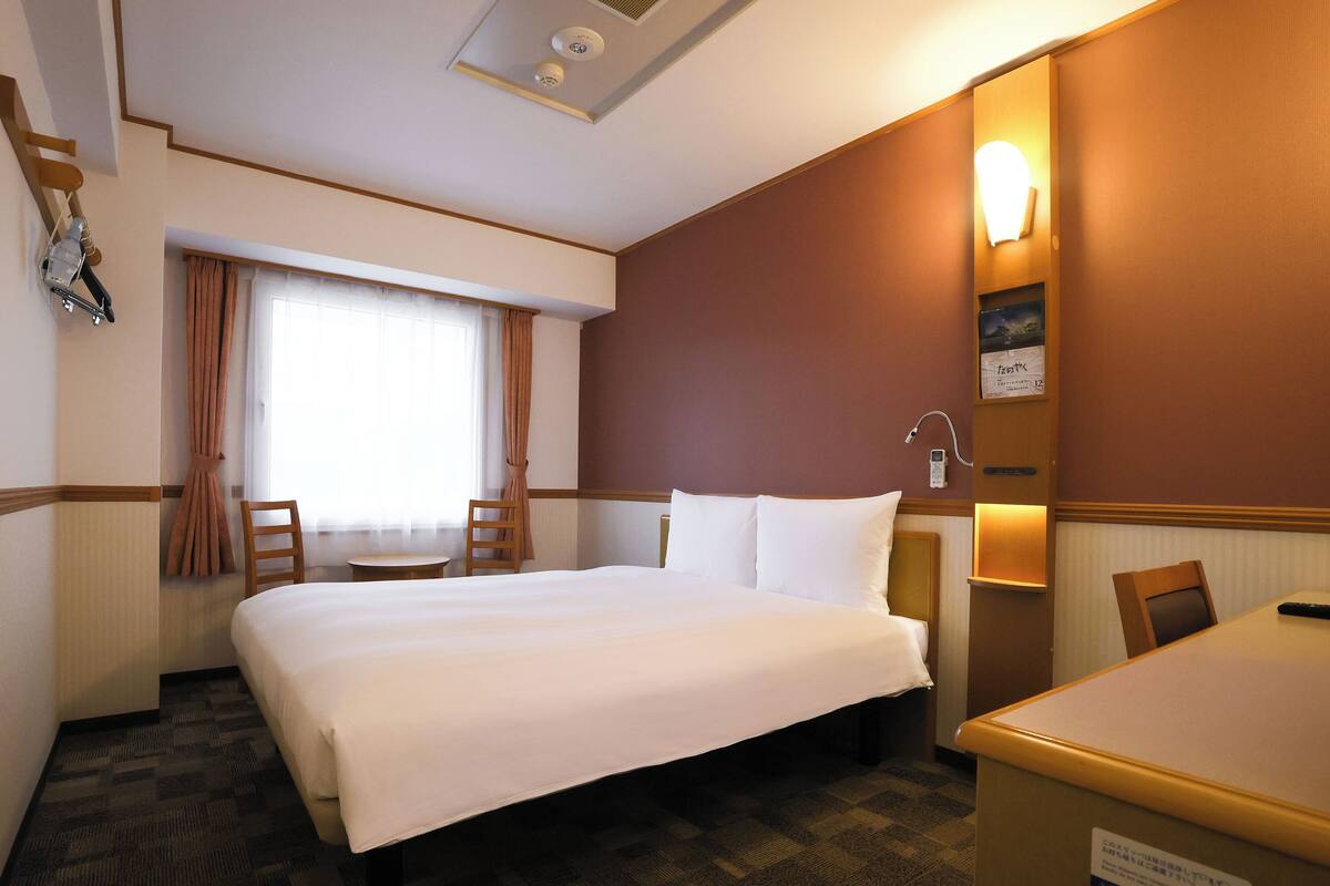 A hotel room with a large bed, a desk, a chair, a window with curtains, and a wall-mounted phone.