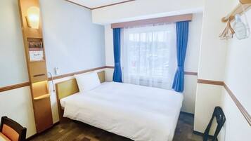 Habitación de hotel con cama, escritorio, silla, ventana con cortinas y estante con un cartel.