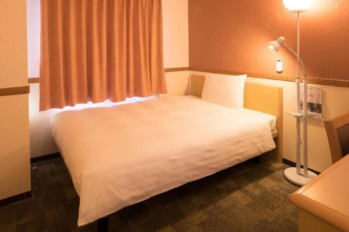 Een hotelkamer met een bed, nachtkastje, lamp en raam met gordijnen.