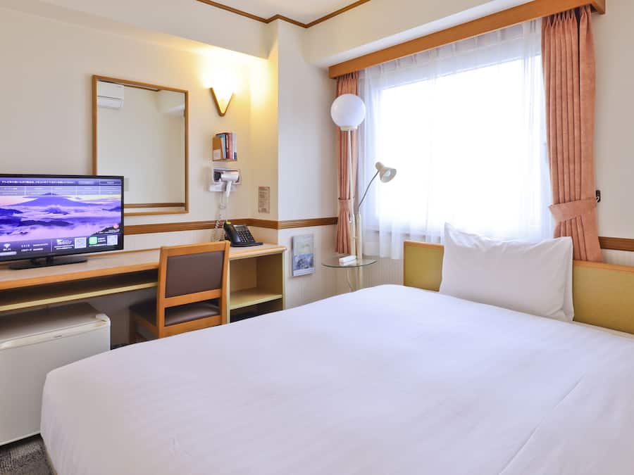A hotel room with a bed, desk, chair, TV, phone, lamp, and window with curtains.