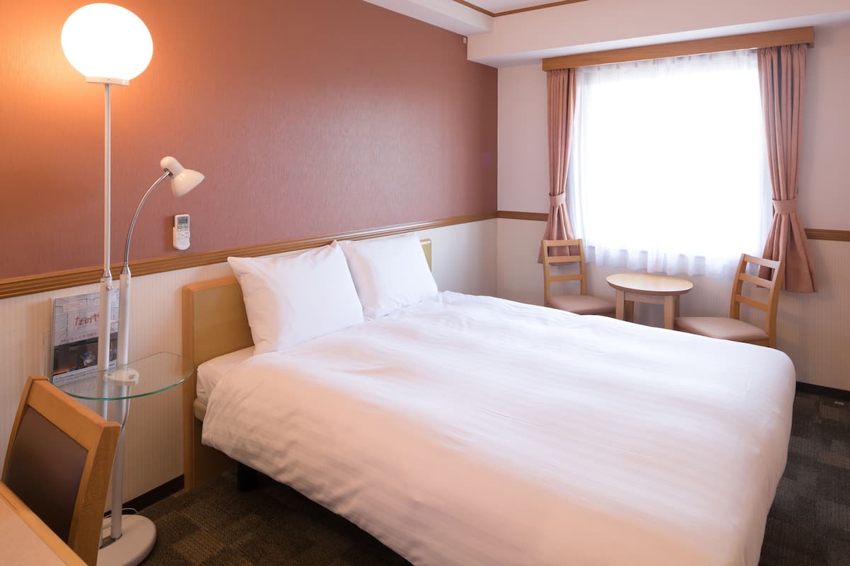 Une chambre d’hôtel avec un grand lit, un bureau avec une lampe, une chaise, une petite table et une fenêtre avec des rideaux.
