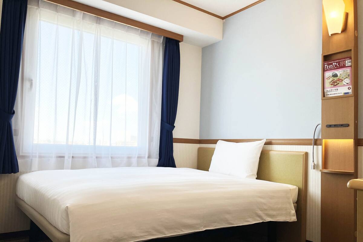Ein Hotelzimmer mit Bett, einem Fenster mit Vorhängen, einem an der Wand befestigten Regal mit einer Zeitschrift und einer Tür.