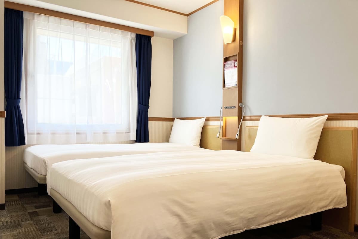 Ein Hotelzimmer mit zwei Betten, einem großen Fenster mit Vorhängen, einer Wandleuchte und einem kleinen Regal mit einem Buch.