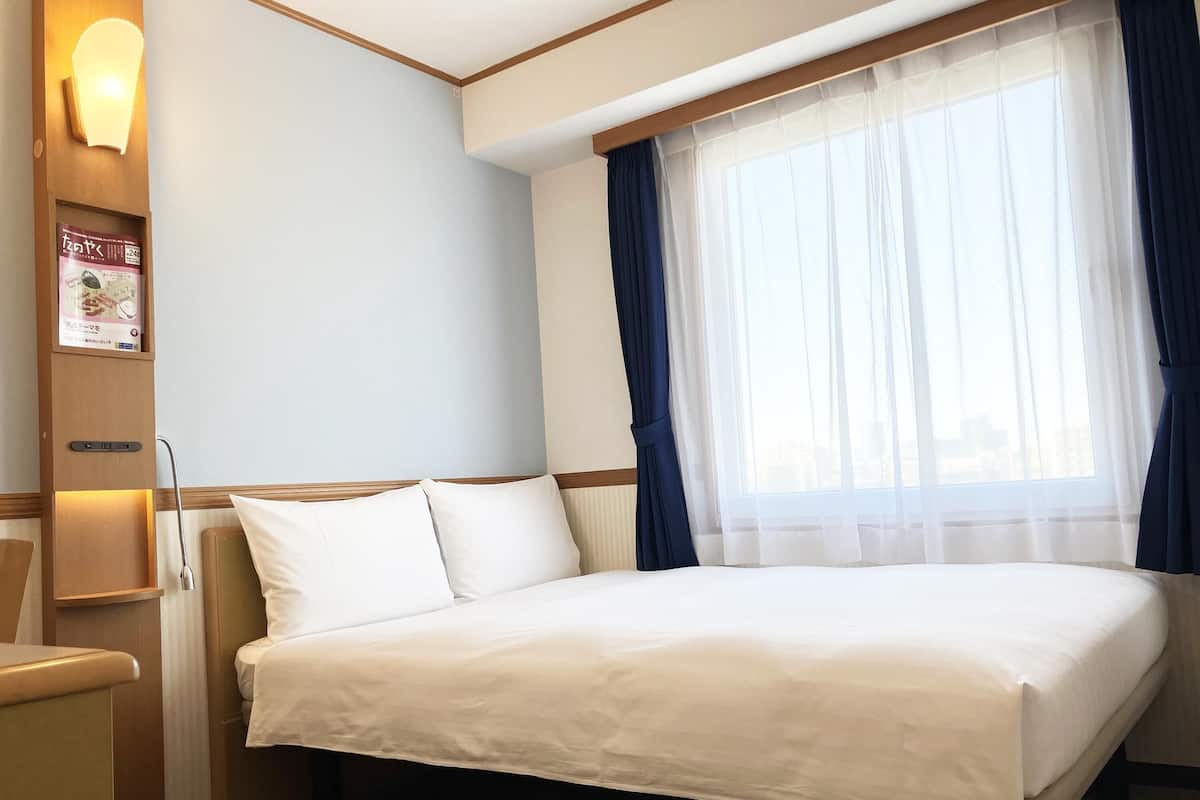 Ein Hotelzimmer mit einem Bett, einem Nachttisch, einer Lampe, einem Fenster mit Vorhängen und einer Wandleuchte.