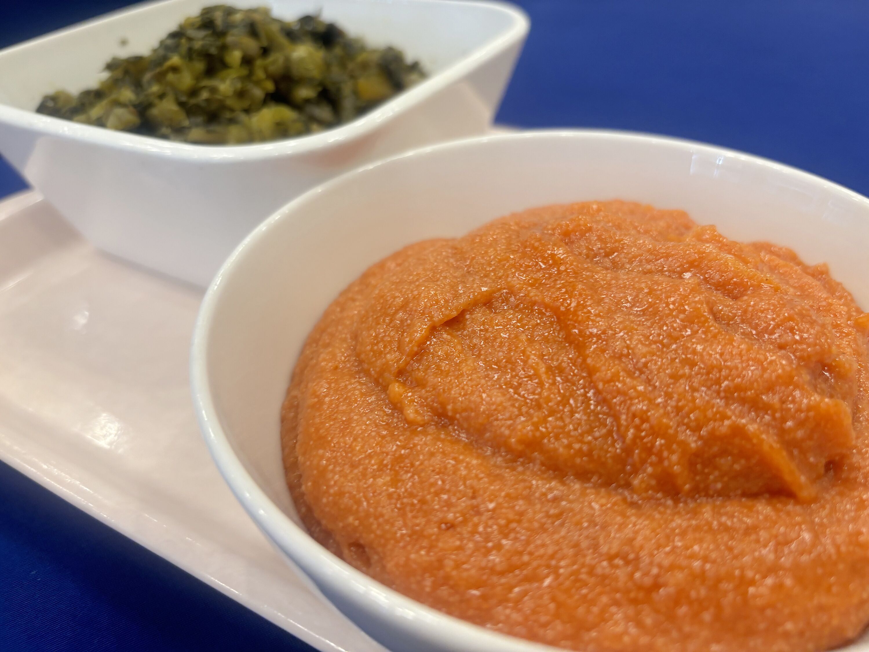 Un tazón blanco con puré de naranja sobre un plato blanco y un mantel azul.