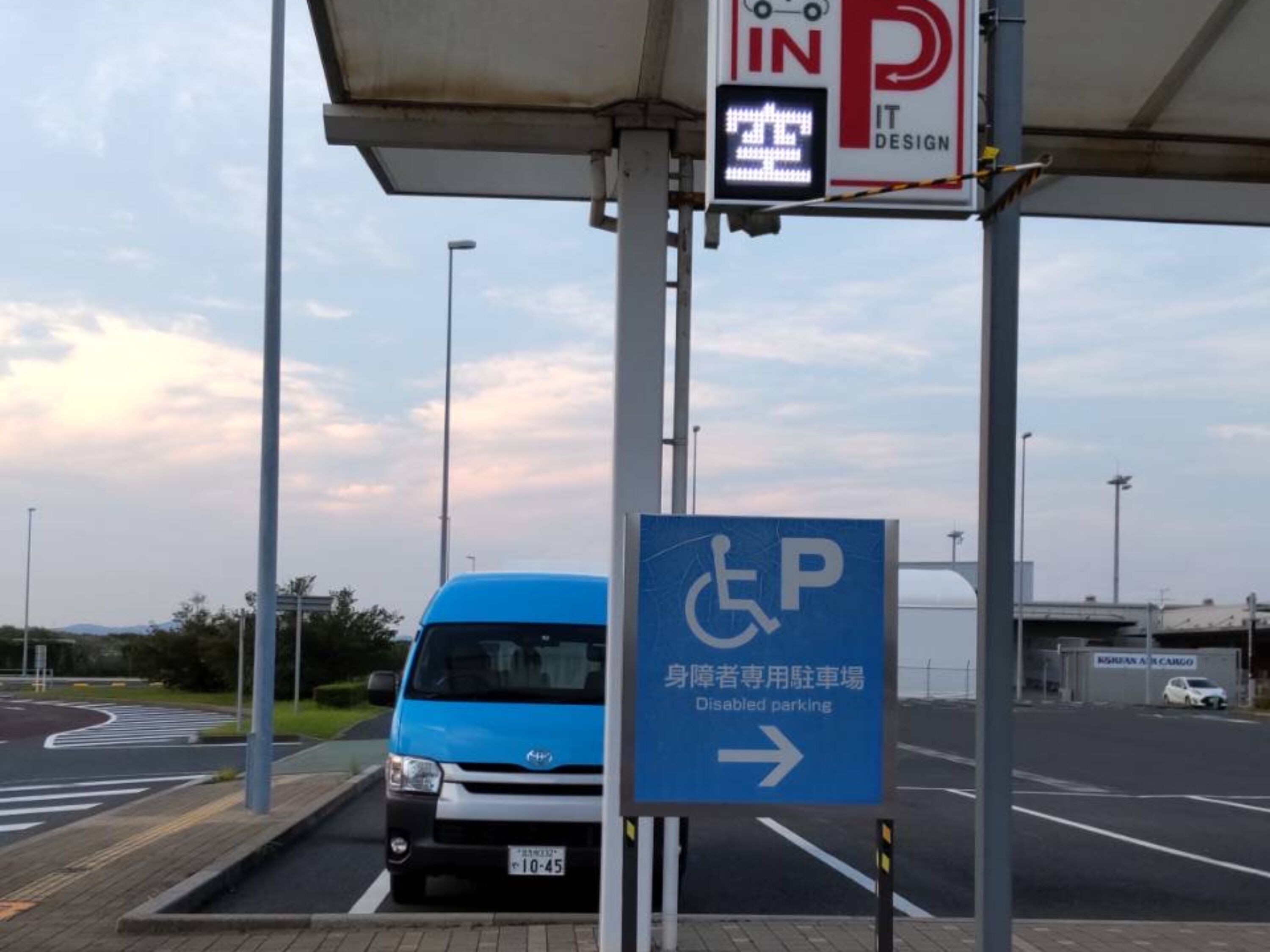A disabled parking sign with an arrow pointing to the right, a blue van parked in front, and a building with a sign that reads "IN IT DESIGN".