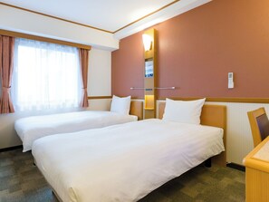 A hotel room with two beds, a large window with curtains, a wall-mounted light, and a small desk.