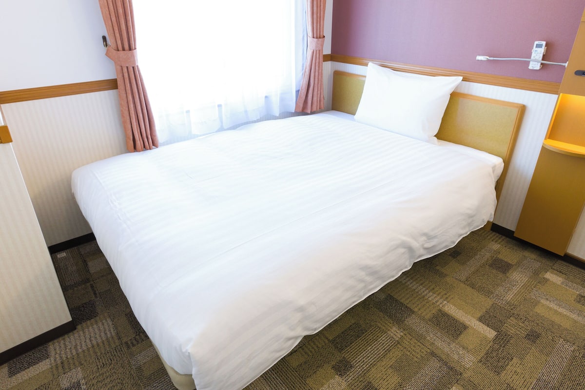 A hotel room with a large bed, a bedside table, a lamp, and a window with curtains.