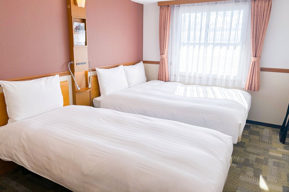 A hotel room with two beds, a nightstand, a lamp, and a window with curtains.