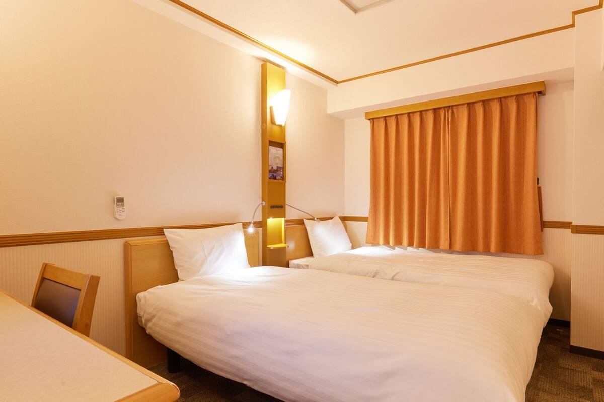 Ett hotellrum med en dubbelsäng, ett skrivbord, ett nattduksbord, en väggmonterad lampa och ett fönster med gardiner.