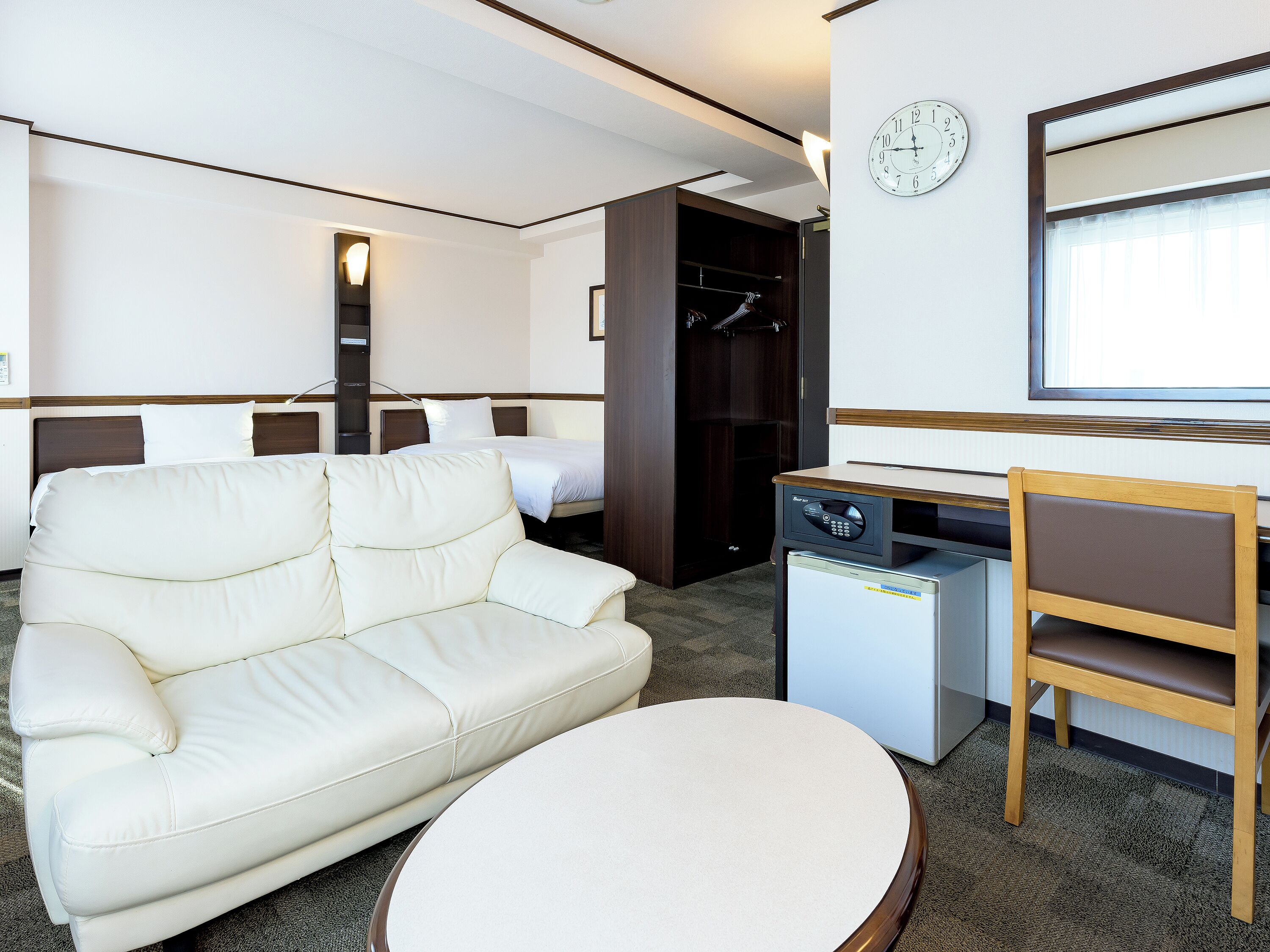 A hotel room with a white sofa, a small table, a desk with a chair, a bed, a wardrobe, and a clock on the wall.