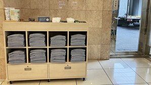 A hotel reception area with a marble wall, a wooden counter with folded towels, and a view of a parked truck outside. - Toyoko Inn Kiryu Station Minami (Kiryu)