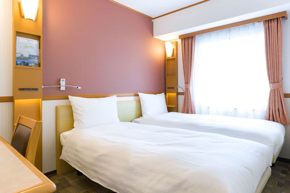 A hotel room with two beds, a desk, a wall-mounted TV, and a window with curtains.