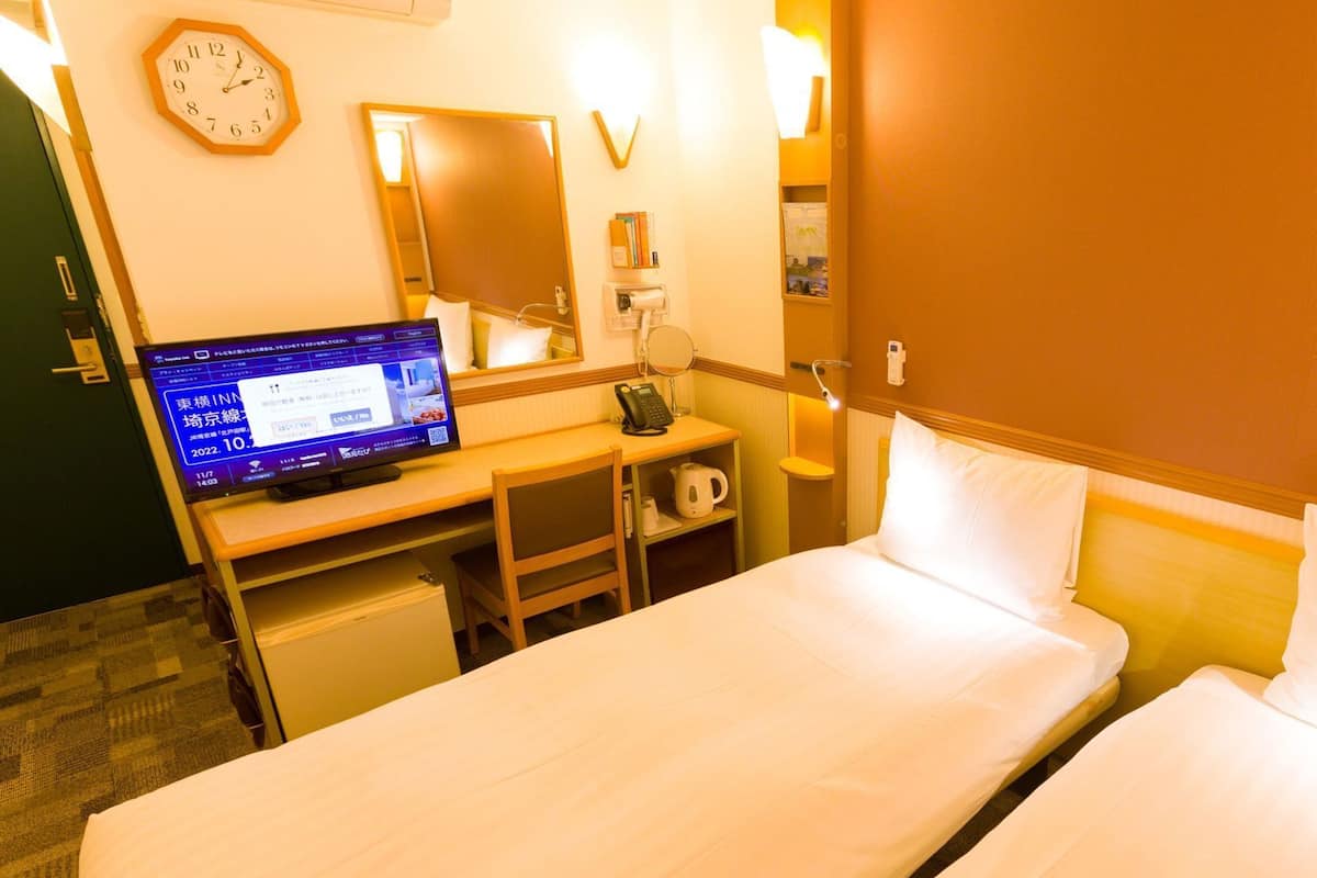 A hotel room with a bed, a desk with a computer, a telephone, a clock, and a mirror.