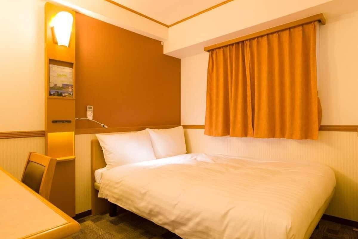 A hotel room with a bed, a desk, a chair, a wall-mounted lamp, and a window with curtains.