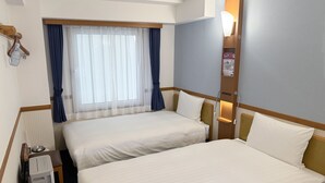 A hotel room with two beds, a window with curtains, a bedside table, a lamp, and a thermostat.